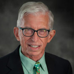 An elderly man with white hair wearing black-rimmed glasses is smiling. He is dressed in a dark suit, green shirt, and a colorful tie. The background is dark and blurred. | Studio 99 Multimedia