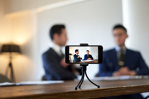 A smartphone on a tripod records two business professionals sitting across each other at a table. They appear to be in a meeting or interview setting, wearing suits and discussing something, with the background out of focus. | Studio 99 Multimedia