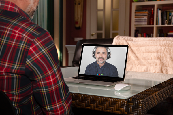 A person wearing a plaid shirt sits at a table, engaging in a video call on a laptop. The laptop screen shows another individual wearing headphones, talking. The setting includes a cozy room with a bookshelf and decorative mask in the background. | Studio 99 Multimedia