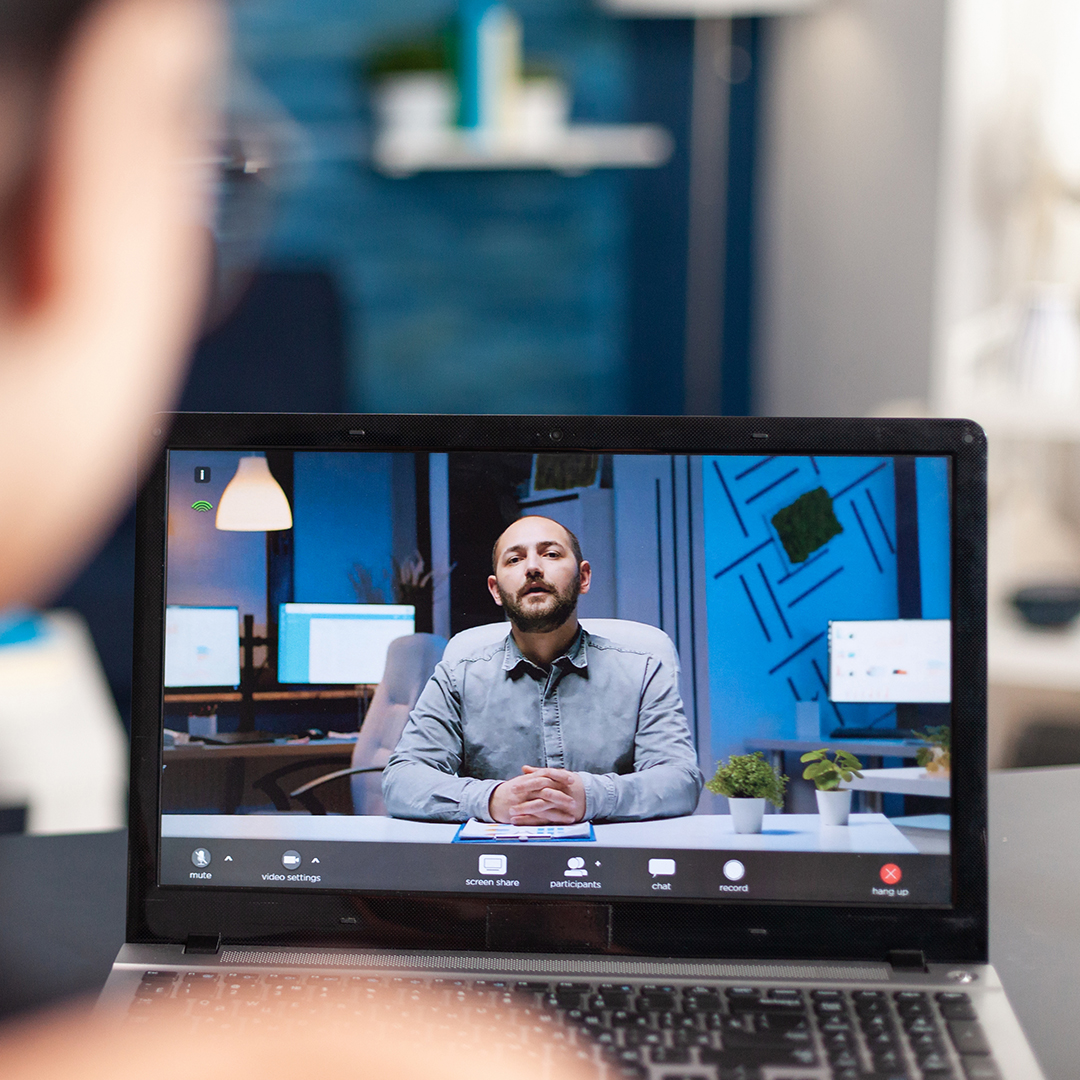 A person is participating in a video call on a laptop. The screen shows a man seated at a desk in a modern office environment, with various office items and computers in the background. The person using the laptop is slightly out of focus in the foreground. | Studio 99 Multimedia