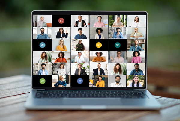 A laptop screen shows a grid of 25 participants in a video conference call. The participants are a mix of men and women, with some displaying their video and others using initials as their display image. The laptop is on a wooden table with a blurred background. | Studio 99 Multimedia