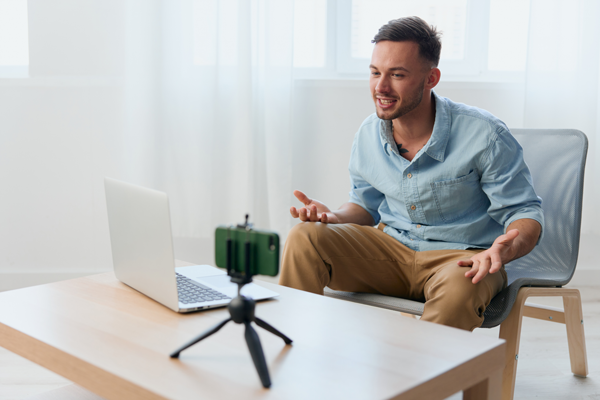 A man sits on a chair in front of a laptop and smartphone mounted on a tripod, engaging in a video call or recording a video. He has a light beard, wears a light blue shirt and tan pants, and appears expressive, gesturing with his hands. | Studio 99 Multimedia