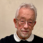 An elderly man with short white hair and wearing glasses is looking at the camera. He is dressed in a black shirt with a white collar and standing against a light-colored wall. | Studio 99 Multimedia