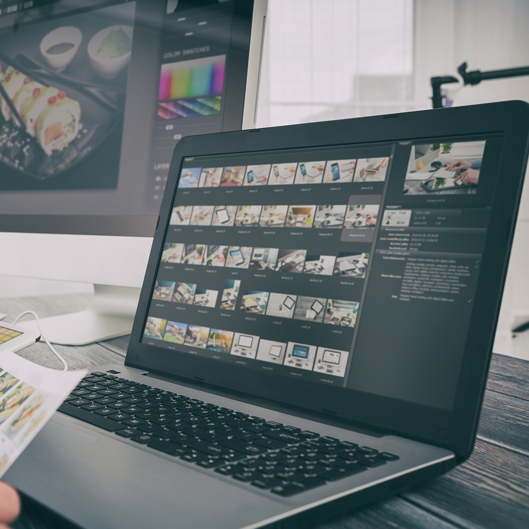 A laptop displaying a photo editing software with numerous thumbnails of images on the screen. Behind it, there is a larger monitor showing a design project. The workspace includes color swatches and various design tools. | Studio 99 Multimedia