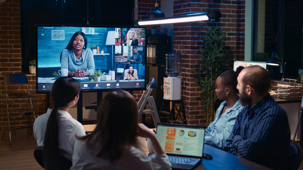 A group of four people sit in an office with exposed brick walls, participating in a video conference call displayed on a large screen. One woman on the screen is speaking, while several others are visible in smaller video windows. A laptop with notes is on the table. | Studio 99 Multimedia