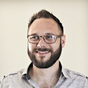 A man with short hair, glasses, and a beard is smiling. He is wearing a light-colored button-up shirt. The background is a plain off-white wall. | Studio 99 Multimedia