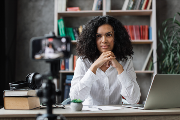 A person with curly hair, wearing a white shirt, sits at a desk with a laptop, books, and headphones, looking at a smartphone on a tripod. Bookshelves and a plant are in the background. | Studio 99 Multimedia