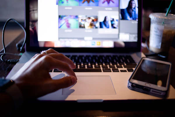 Close-up of a person using a laptop with a photo editing software open. Their hand is on the trackpad, and a smartphone lies next to the laptop on the right. In the background, an iced drink with a straw sits on the table. | Studio 99 Multimedia