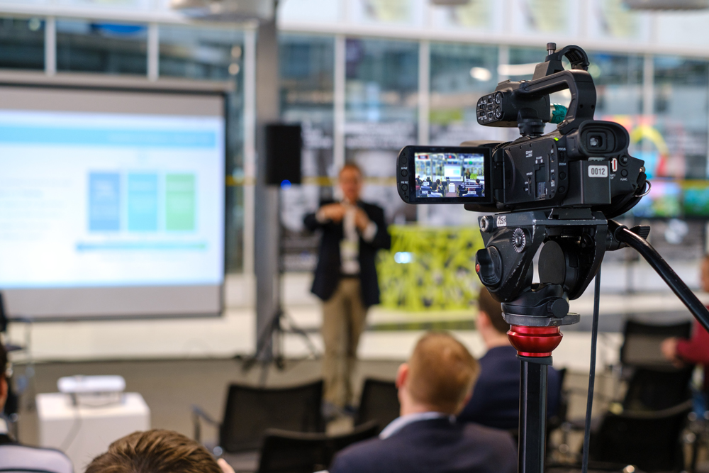 A video camera on a tripod records a speaker giving a presentation in a modern conference room. The speaker is slightly blurred in the background, and a screen displaying slides is visible. Several attendees are seated in the foreground. | Studio 99 Multimedia