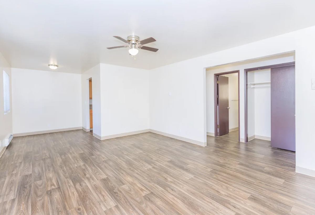 Spacious empty room with light-colored walls, wood-style flooring, a ceiling fan, open closet, and doorways leading to other rooms. Natural light comes from windows on the left. | Studio 99 Multimedia