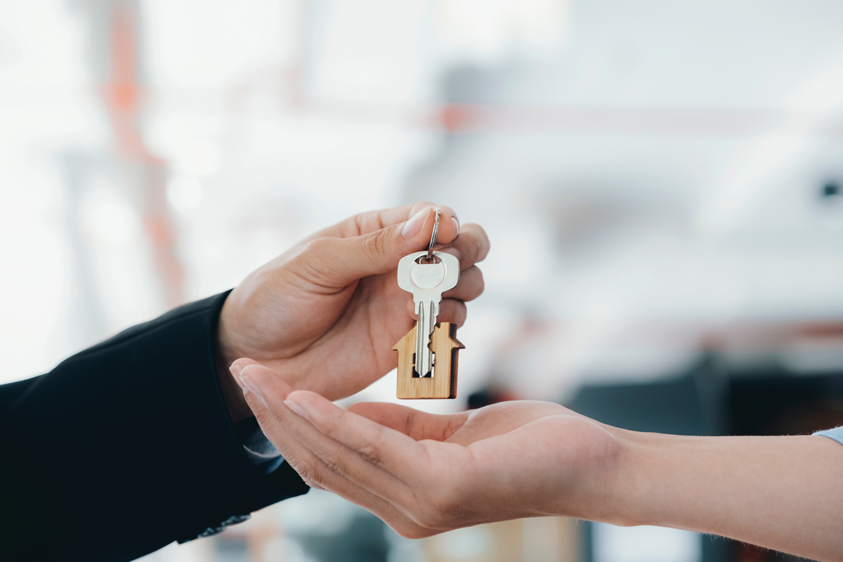 A person hands over a key attached to a house-shaped keychain to another person, symbolizing buying, selling, or renting a home. The background is blurred, focusing on the exchange. | Studio 99 Multimedia